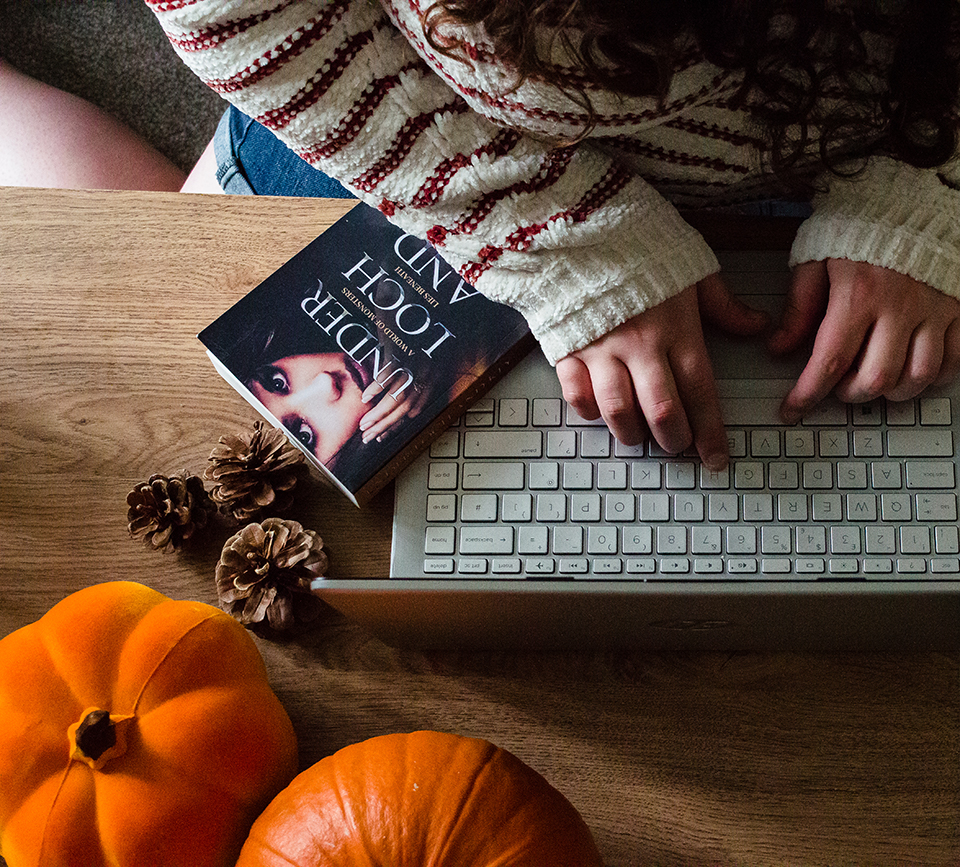An image of some pumpkins, a book, keyboard and hands on top of a desk.